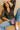 Woman sitting on a couch holding a purple handbag, wearing a patterned top and jeans.