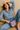 Woman sitting on a couch holding a purple handbag, wearing a blue floral shirt and jeans.
