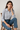 Woman sitting on a wooden chair wearing a light blue checkered shirt and blue jeans.