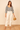 Woman wearing a black and white polka dot blouse and white pants, holding a straw bag on a beige background