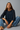 Woman wearing a navy blue top and blue jeans sitting on a gray background
