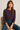 Woman wearing a dark purple long-sleeve top and blue jeans sitting on a chair.